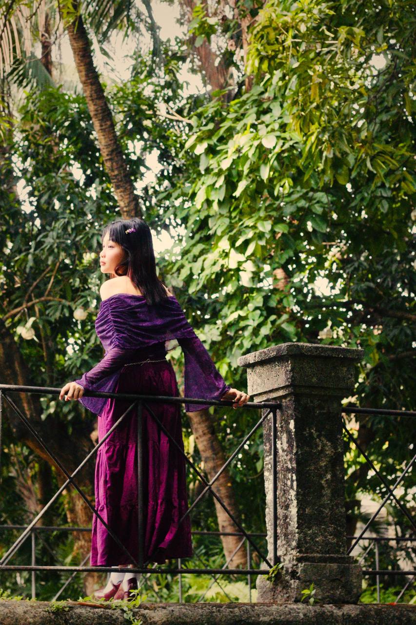Daena Amor in a purple off-shoulder dress, standing at an iron railing and looking out into a garden of trees