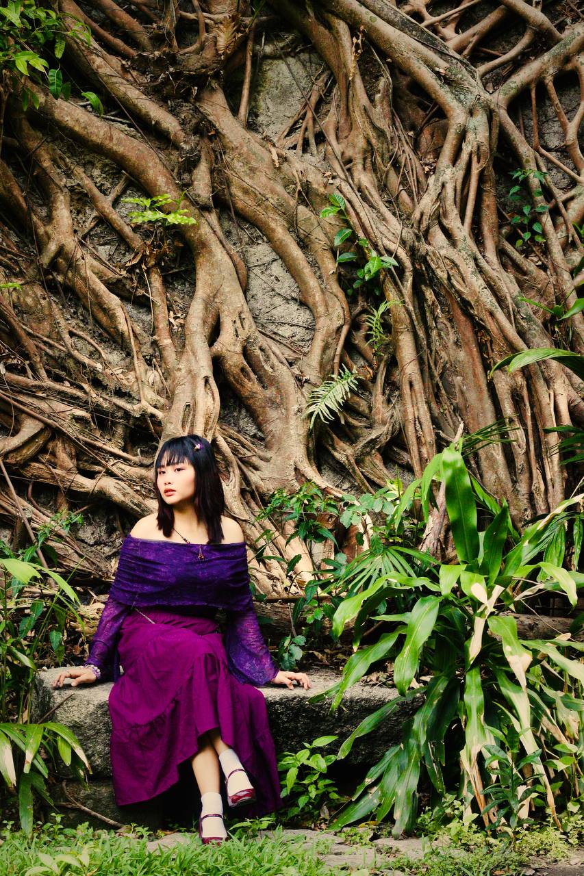Daena Amor seated on a stone ledge in front of a wall of banyan roots, surrounded by ferns
