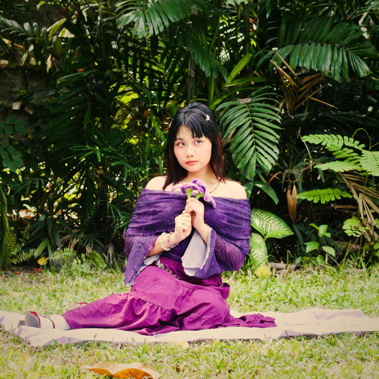 Daena Amor kneeling on a cloth spread over grass, holding a purple flower to her chest